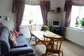 A living room with a sofa, a wooden dining table, and two chairs. There is a television on a stand, a lamp, and a window with curtains. - Holiday apartment with 2 bedrooms (Bad Gottleuba-Berggießhübel)