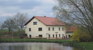 A two-story building with a red roof, situated next to a body of water, surrounded by trees and a fence. - Holiday apartment with 2 bedrooms (Bad Gottleuba-Berggießhübel)