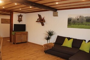 Ein Wohnzimmer mit Holzboden, einem braunen Sofa mit grünen Kissen, einem Fernseher auf einem Holzschrank, einer an der Wand hängenden Uhr und einem gerahmten Landschaftsbild.