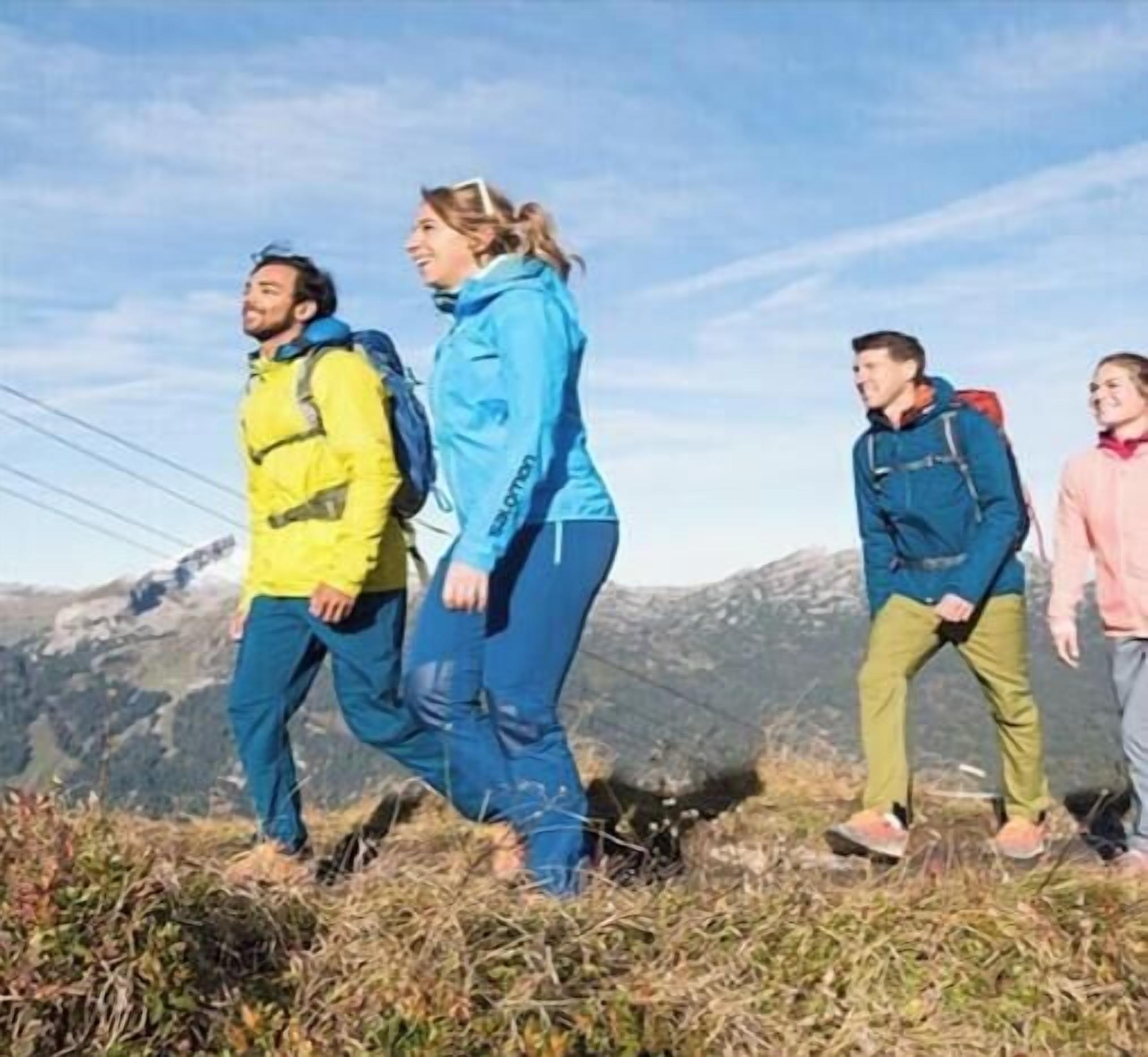 Quatre personnes font de la randonnée sur un sentier de montagne herbeux, avec une chaîne de montagnes en arrière-plan.