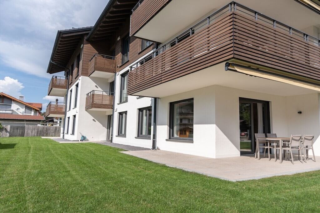 A modern two-story residential building with a balcony, a patio with outdoor furniture, and a well-maintained lawn.