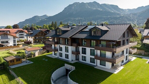 Um edifício residencial moderno, com vários andares, teto plano, grandes janelas e um gramado bem cuidado numa área montanhosa.