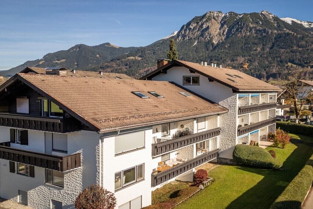 Un edificio moderno de dos pisos con balcones, fachada de piedra y montañas al fondo.