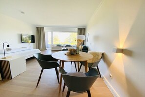 A modern living room with a sofa, a dining table with chairs, a flat-screen TV mounted on the wall, and a window with curtains. - Holiday apartment with 1 bedroom (Travemünde)