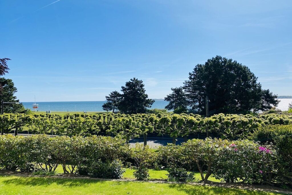 Ein angelegter Garten mit verschiedenen Sträuchern und Bäumen, ein klarer, blauer Himmel und ein Gewässer in der Ferne.