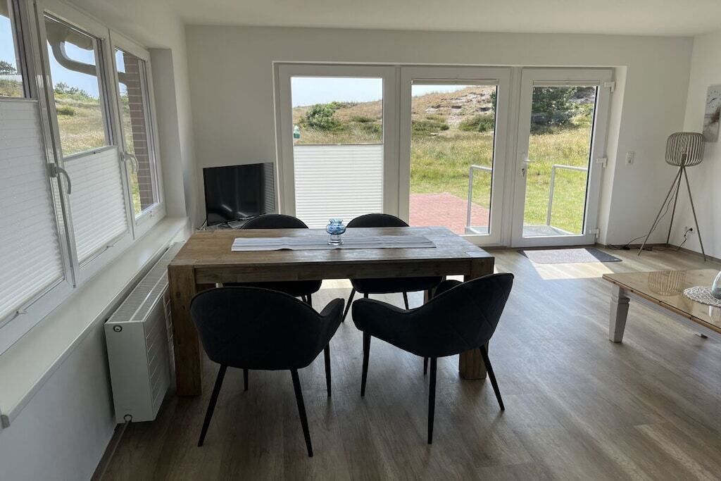 Une pièce avec une table en bois, deux chaises noires, un téléviseur et une vue sur un espace extérieur, accessible par de grandes portes vitrées.