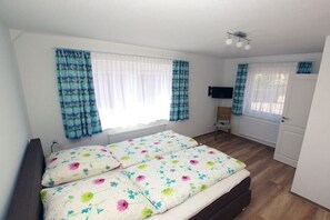 A bedroom with a bed, curtains, a television, and a door. - Apartment 2 in the Mare Cimbria building (Norddorf auf Amrum)