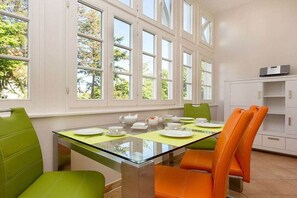 A dining area with a glass table, green and orange chairs, white cabinets, and large windows with a view of trees. - Havana at Villa Malepartus (Binz)