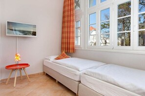Ein Zimmer mit zwei Einzelbetten, einem kleinen Tisch mit einer Lampe, einem Bild an der Wand und einem Fenster mit Vorhängen.