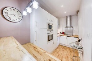 A compact kitchen with white cabinetry, a wooden countertop, a built-in oven, a microwave, a stainless steel range hood, and a wall-mounted clock.