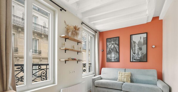 Ein Zimmer mit einer Couch, einem Heizkörper, einem Fenster mit Blick auf ein Gebäude und zwei gerahmten Bildern an der Wand.
