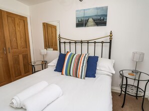 A neatly made bed with white linens and striped pillows, a wooden wardrobe, a small round table with a lamp, and a painting of a pier on the wall. - Ty'r Heulwen (Aberdovey)