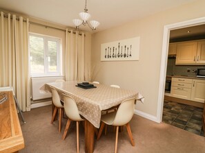 A dining area with a table, chairs, a chandelier, and a kitchen visible through an open door. - Ty'r Heulwen (Aberdovey)