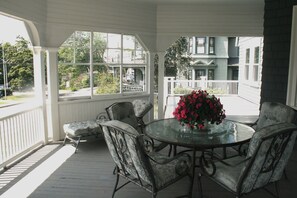 A covered porch with a round table and six chairs, a potted plant with flowers, and a view of a street and buildings. - Large 14bed Family Retreat a Stroll to York Harbor Beach and Venues (York)