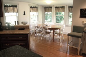 A kitchen with a wooden table and chairs, a granite countertop, and a view of the outdoors through large windows. - Large 14bed Family Retreat a Stroll to York Harbor Beach and Venues (York)
