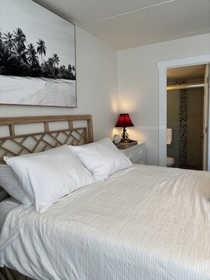 Habitación de hotel con una cama, una mesita de noche con una lámpara, un cuadro de una playa y una puerta que da al baño.