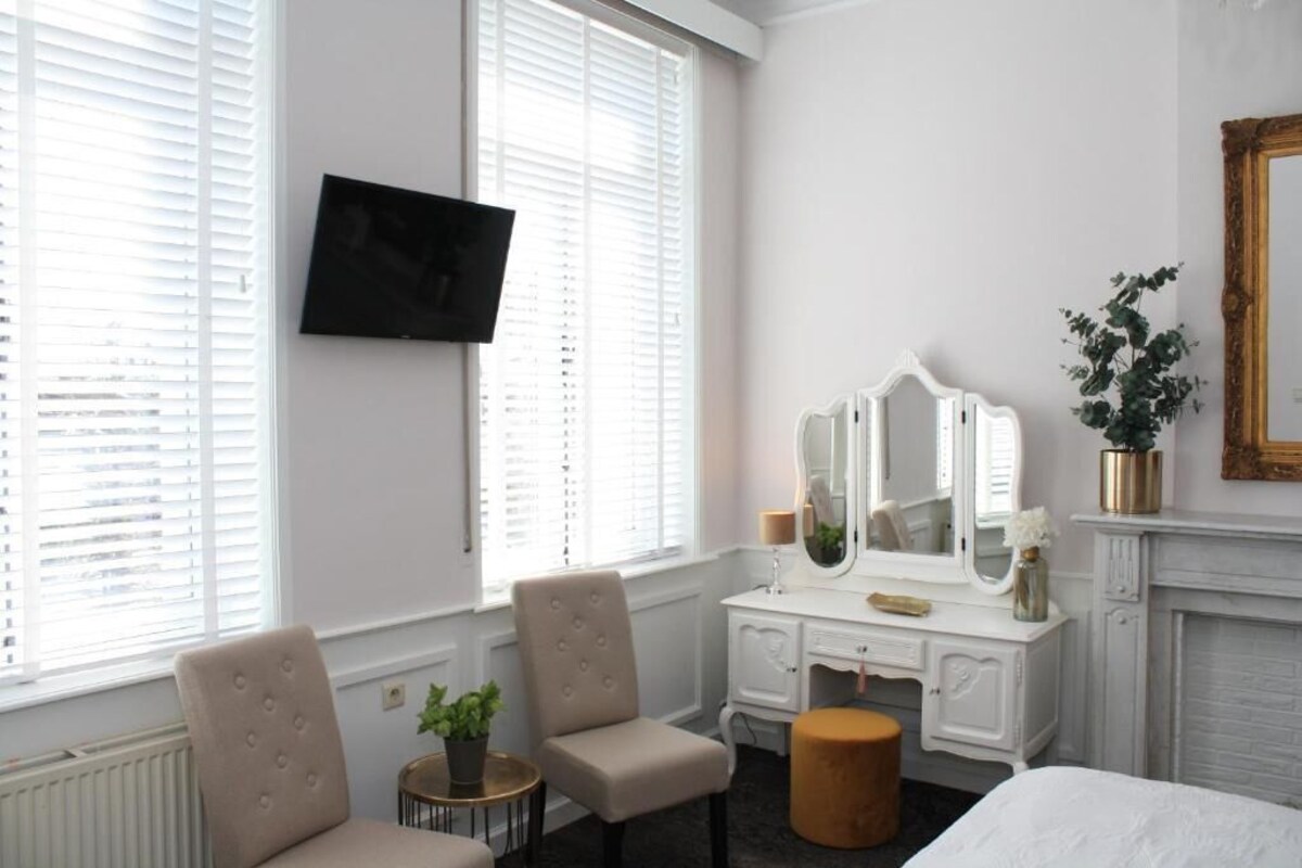 Une chambre d’hôtel comprenant un lit, une coiffeuse avec un miroir, deux chaises, une petite table ronde et une plante en pot.