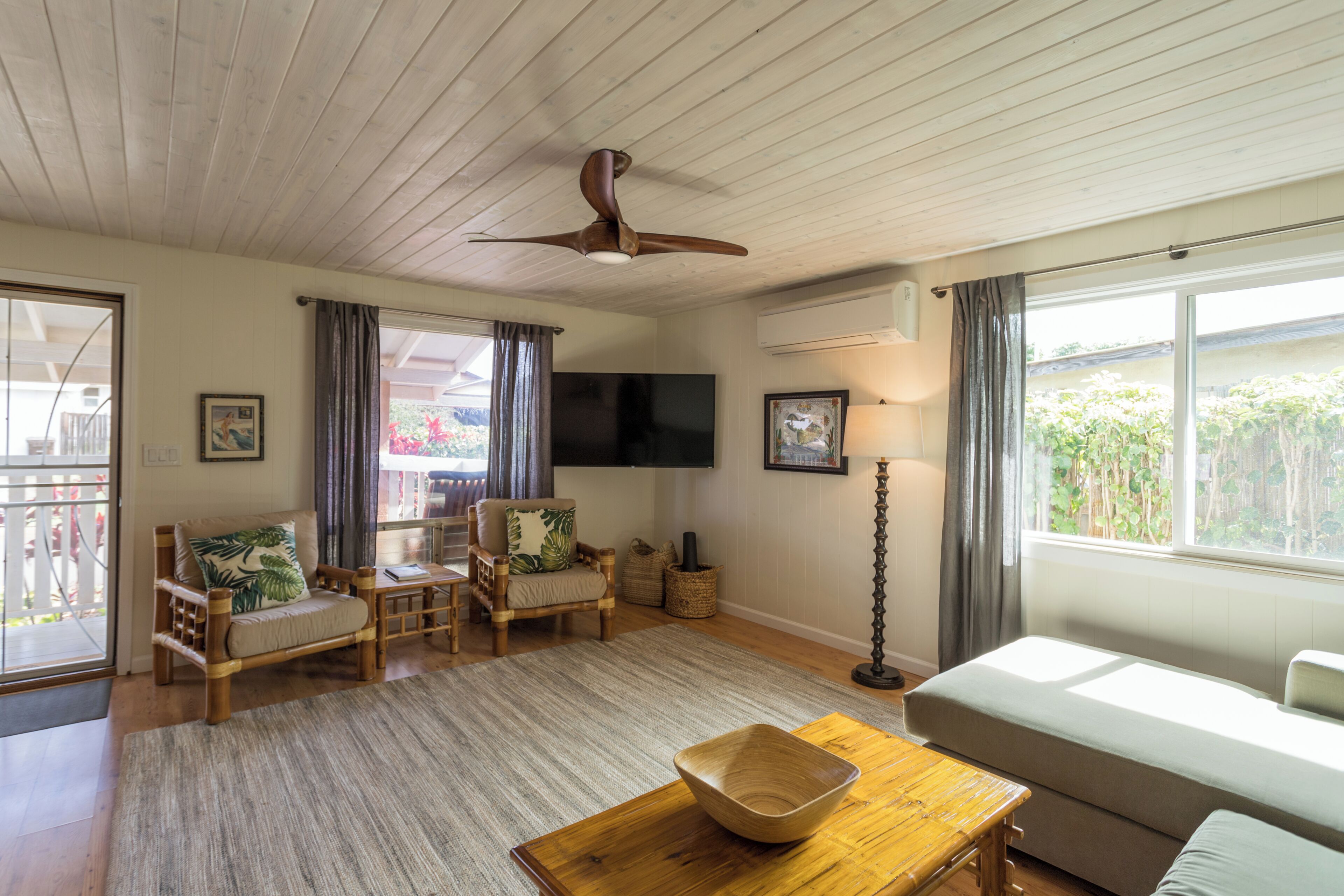 Una sala de estar con ventilador de techo, televisor de pantalla plana, sofá, mesa de centro y ventana con cortinas.