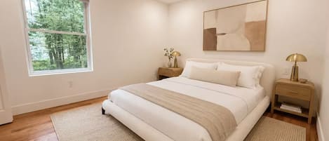 A bedroom with a large bed, a nightstand with a lamp, a window with a view of trees, and a wall-mounted abstract art piece.