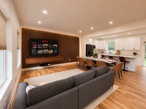 A modern living room with a grey sofa, wooden flooring, a dining area with a table and chairs, and a kitchen with white cabinets. - New Luxury Stay with Private Pool, Hot Tub, Sauna (Tobyhanna)