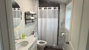 Une salle de bain avec un lavabo, des toilettes et un rideau de douche, tous de couleur blanche. Un miroir est placé au-dessus du lavabo et un porte-serviettes se trouve à côté.