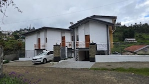 A two-story residential building with a white exterior, a car parked in front, and a hilly landscape in the background.