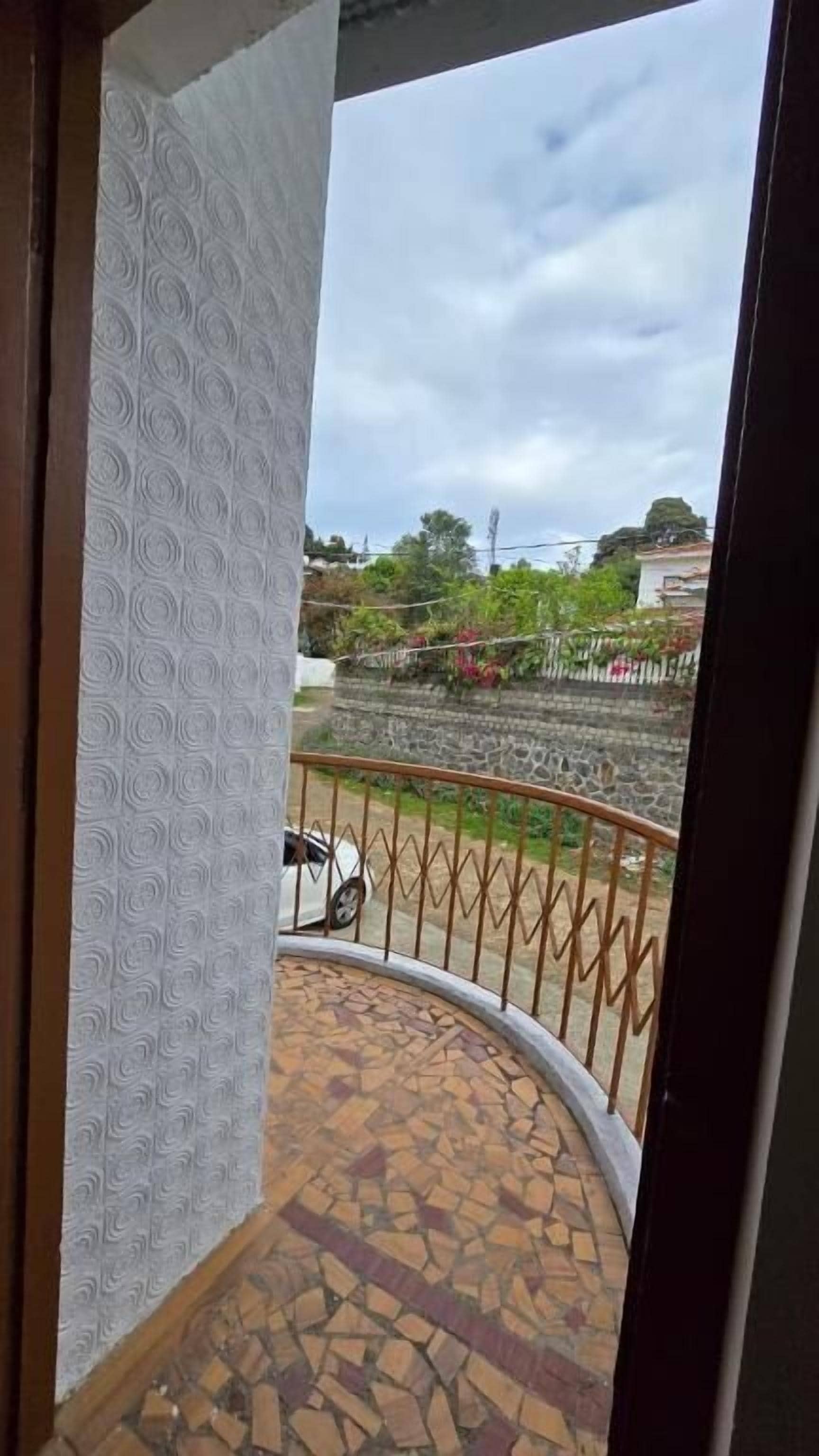 A balcony with a patterned wall, a tiled floor, and a view of a garden and buildings in the distance.