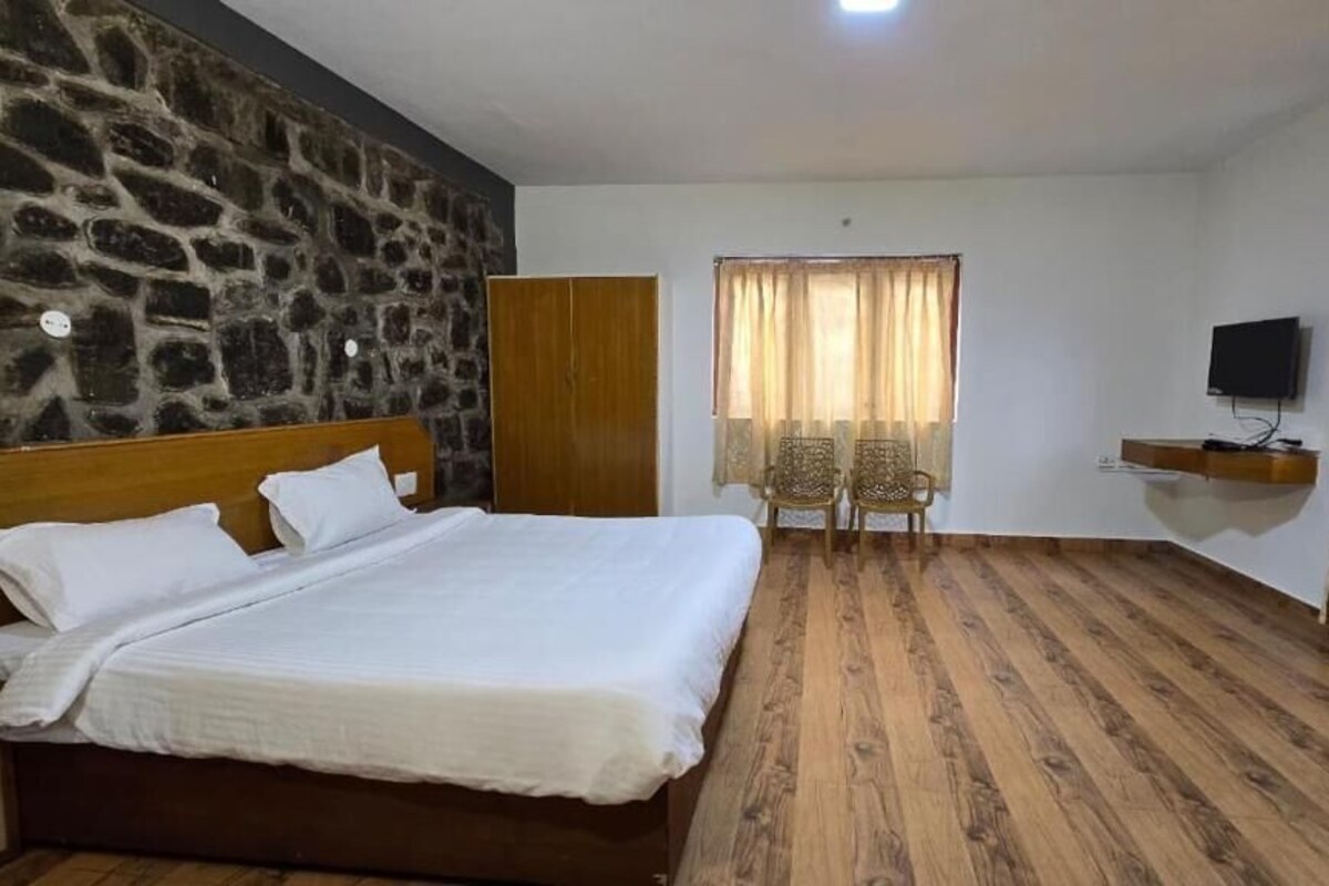 A bedroom with a large bed, a stone wall, a wooden wardrobe, a television on a shelf, and a window with curtains.