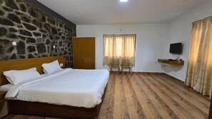 A bedroom with a large bed, a stone wall, a wooden wardrobe, a television on a shelf, and a window with curtains. - Vedha Holidays (Kodaikanal)
