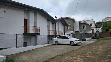 Bairro residencial com um carro branco estacionado na entrada de uma casa, casas com varandas e uma cerca.