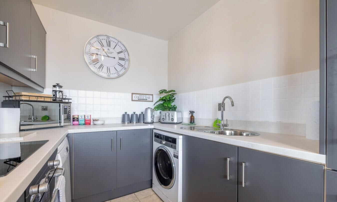 Een moderne keuken met grijze kastjes, een ingebouwde wasmachine, een bord met 'coffee bar' en een wandklok.