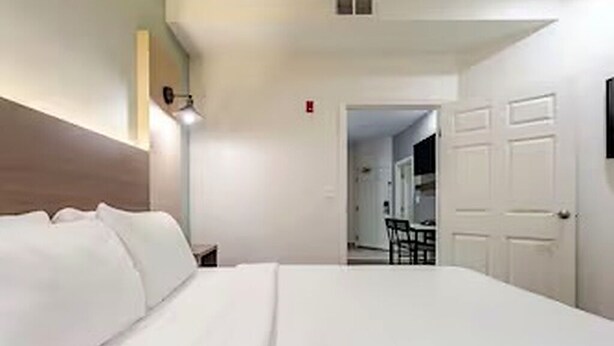 A hotel room with a large bed, a bedside table, a lamp, and a door leading to a dining area.