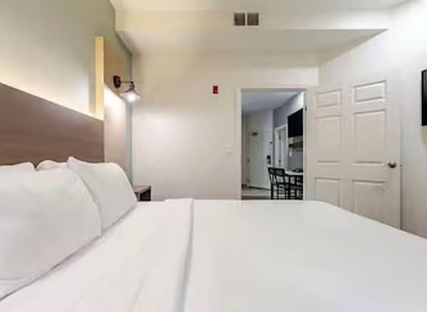 A hotel room with a large bed, a bedside table, a lamp, and a door leading to a dining area.