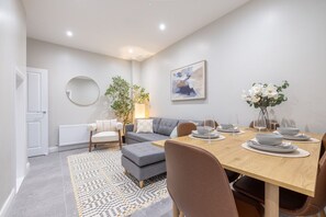 Ein modernes Wohnzimmer mit einem grauen Sofa, einem runden Spiegel, einem Esstisch gedeckt mit Tellern und Gläsern sowie einem Gemälde an der Wand.