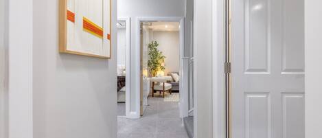 Un pasillo con una puerta blanca a la derecha, una obra de arte enmarcada a la izquierda y un comedor con mesa y sillas al fondo.