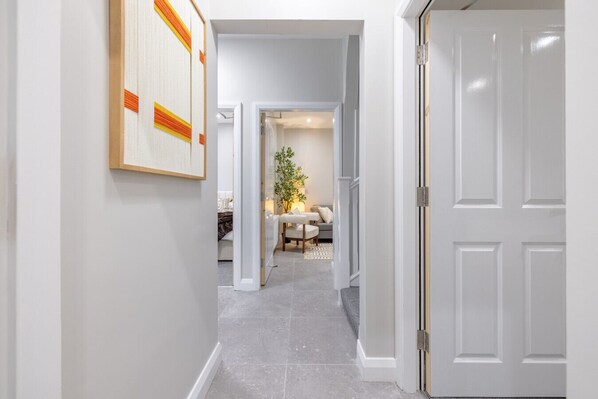 Un couloir avec une porte blanche sur la droite, une œuvre d’art encadrée sur la gauche, et un coin repas avec une table et des chaises à l’arrière-plan.