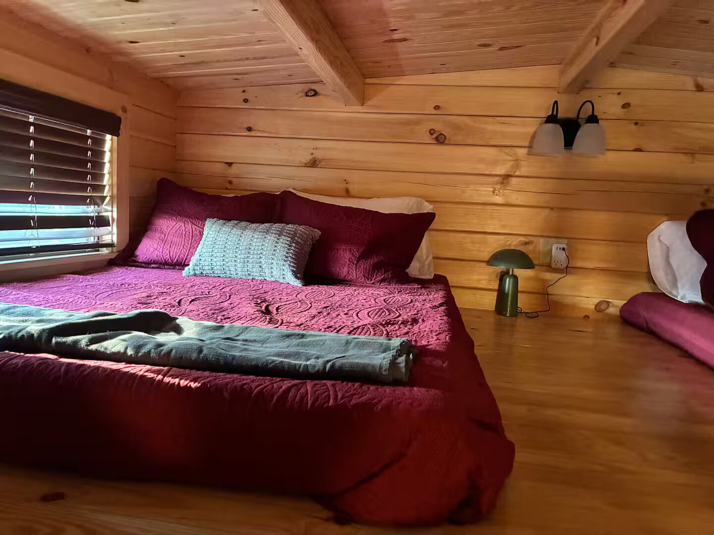 Una habitación tipo cabaña de madera con cama, almohadas, una lámpara y una ventana con persianas.