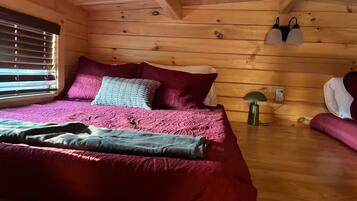 Una habitación tipo cabaña de madera con cama, almohadas, una lámpara y una ventana con persianas.