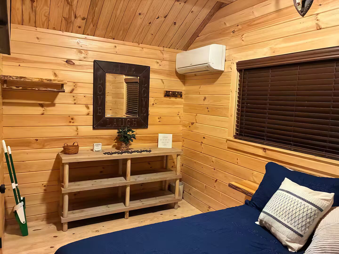 A wooden cabin interior with a bed, a mirror, a shelf with a plant, and a window with blinds.