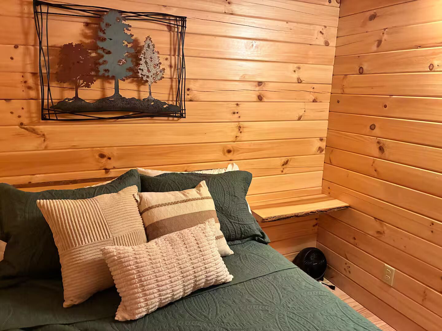 A cozy room with wooden walls, a bed with green bedding and multiple pillows, a small shelf, and a wall-mounted picture of trees.