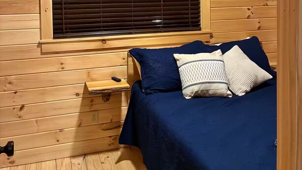 A wooden cabin interior with a bed, a window with blinds, a mounted deer head, and an exit sign.