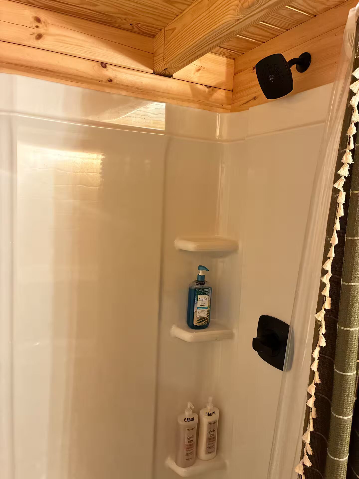Zona de ducha con techo de madera, un cabezal de ducha negro y dos botellas de jabón corporal Cabin Suave.