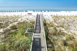Eine Holzpromenade führt zu einem Strand mit weißem Sand und blauen Sonnenschirmen.