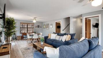 Ein modernes Wohnzimmer mit einem blauen Sofa, einem Esstisch mit Stühlen, einem Deckenventilator und einer Treppe.