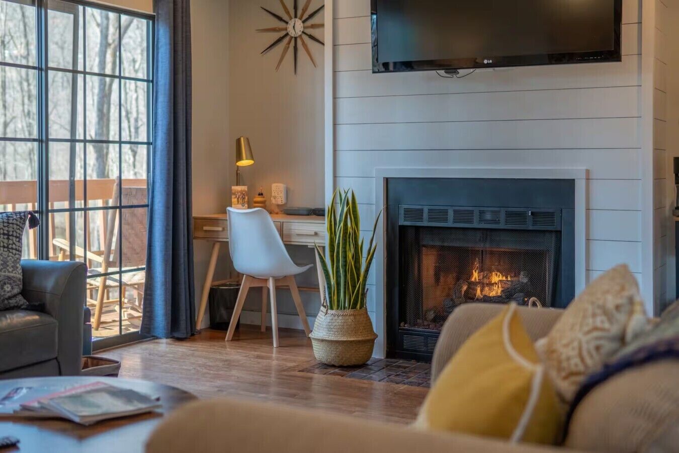 A living room with a fireplace, a television mounted above it, and a view of the outdoors through large windows.
