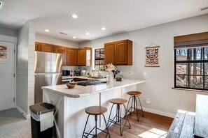 Una cocina moderna con armarios de madera, un refrigerador de acero inoxidable, una encimera de mármol y un amplio ventanal con vistas a árboles.