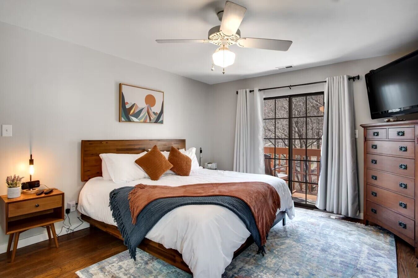 A bedroom with a bed, a nightstand, a dresser, a ceiling fan, a painting, and a window with curtains.