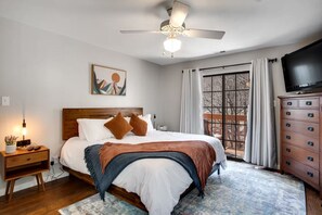 Un dormitorio con cama, mesita de noche, cómoda, ventilador de techo, un cuadro y ventana con cortinas.