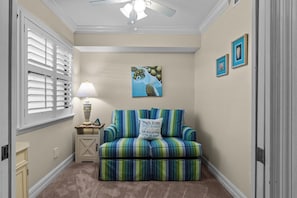 Ein Zimmer mit Deckenventilator, einem Fenster mit Jalousien, einer Lampe, einem gestreiften Sofa mit Kissen und einem Bild an der Wand.