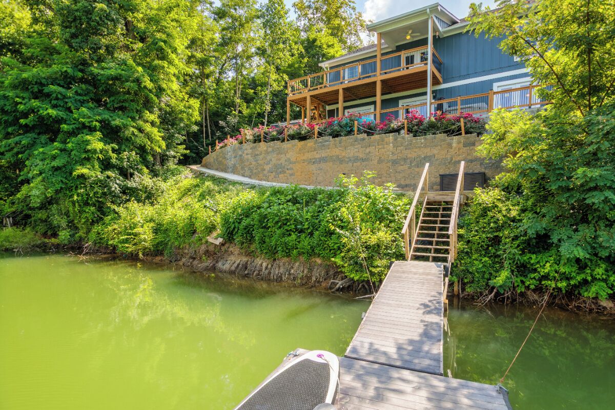 A wooden dock leading to a house with a balcony, surrounded by lush greenery and a calm body of water.